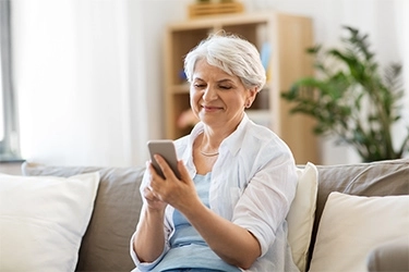 A woman with grey hair smiling at her phone on a sofa