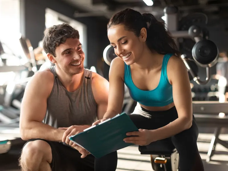 A trainer and client smiling as they review a workout plan on a clipboard in the gym