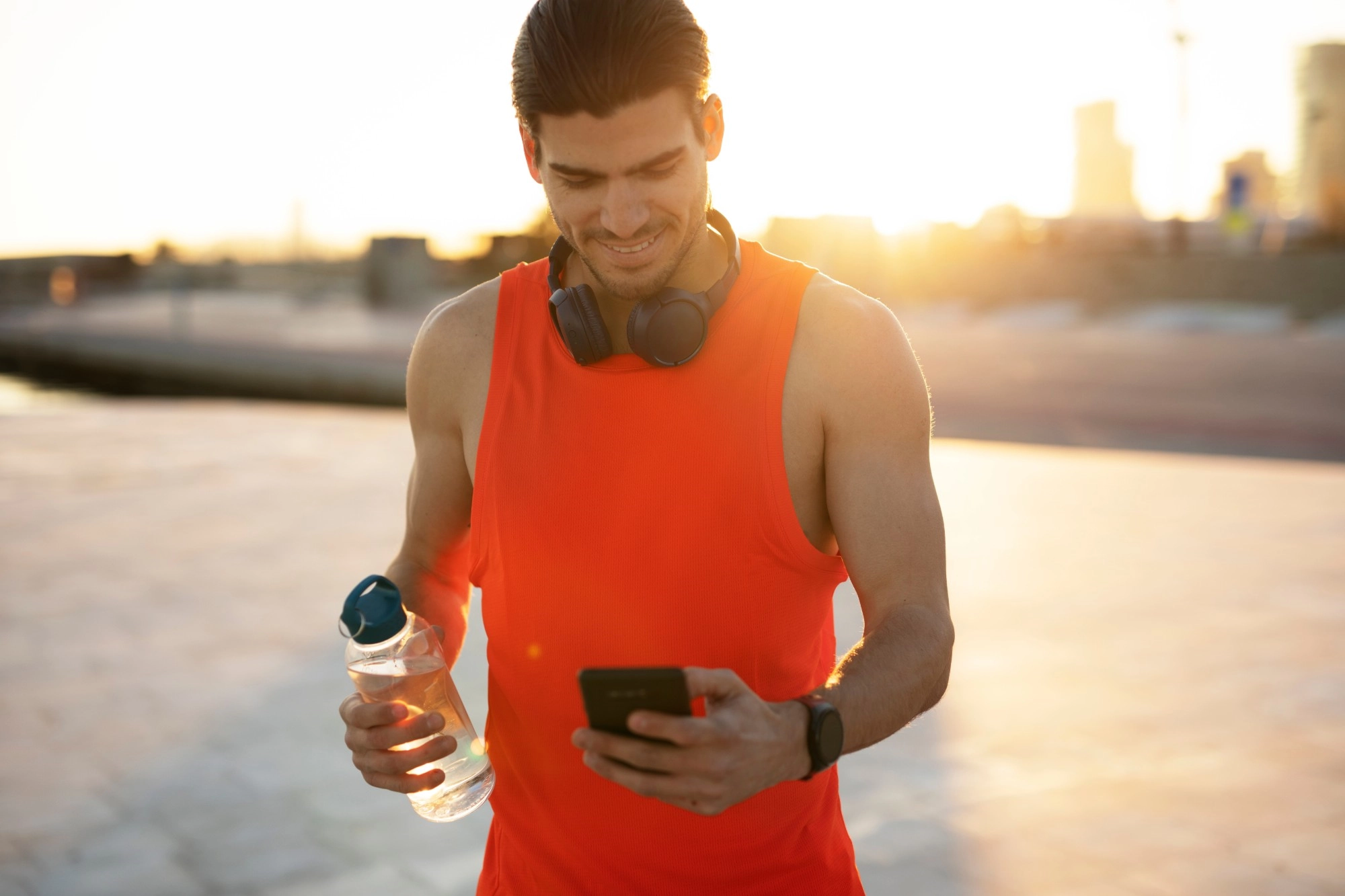 A smiling man holding a water bottle and checking his phone at sunset