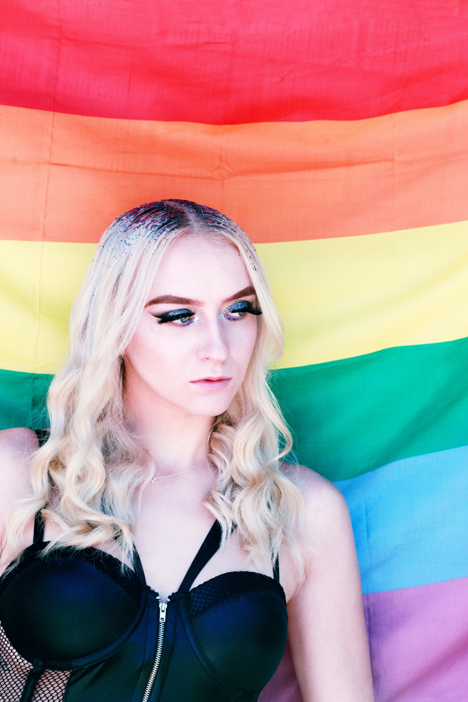 Persona rubia con un top negro delante de una bandera arcoíris