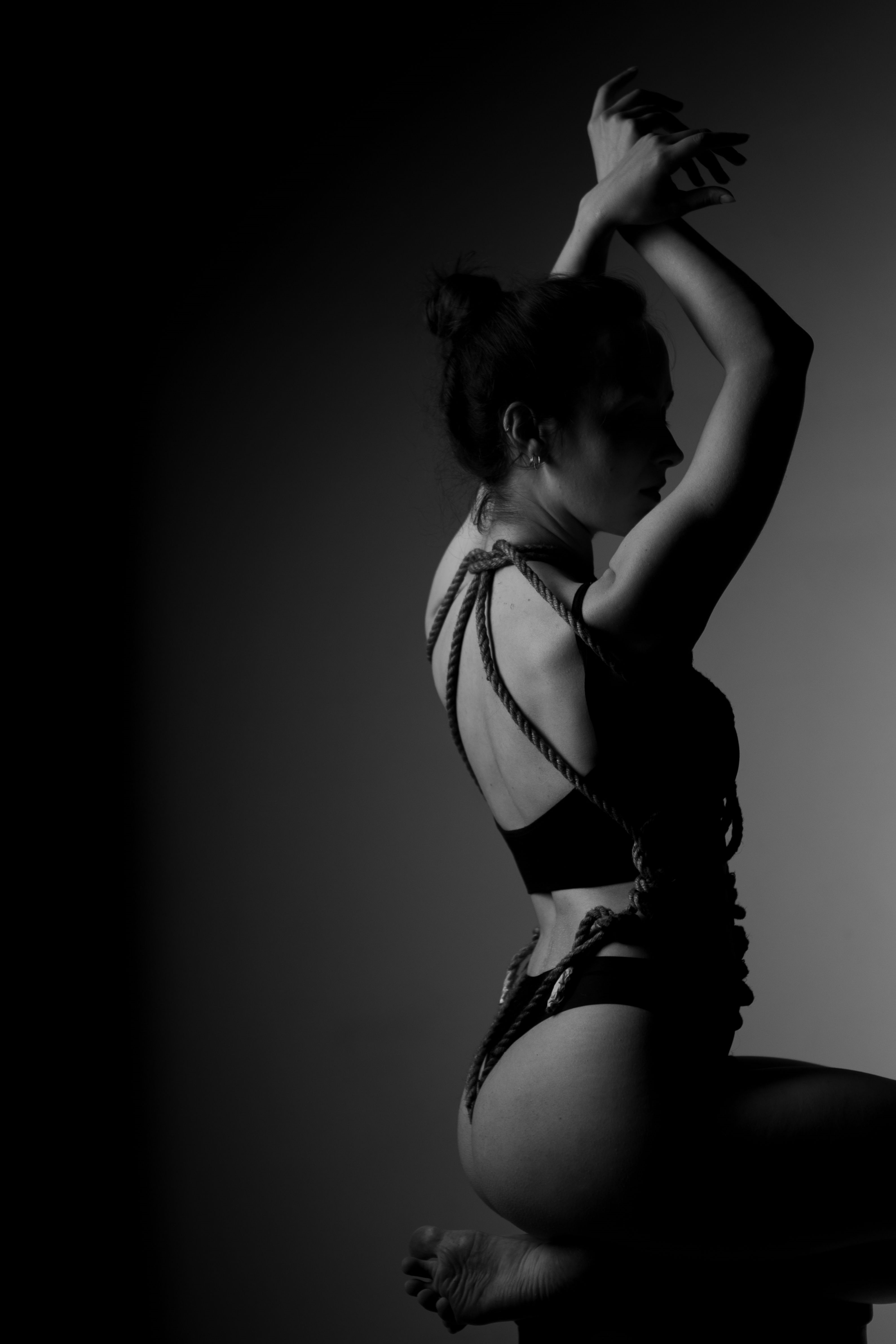 Black-and-white profile portrait of a woman with shibari rope tied across her bare back and shoulders, arms raised