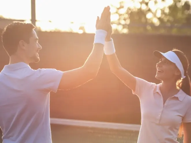 A man and woman in tennis whites high-fiving on court at sunset