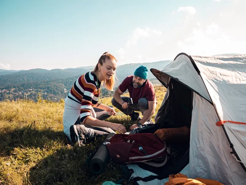 A woman and man pitching a tent on a sunny hillside with mountains behind