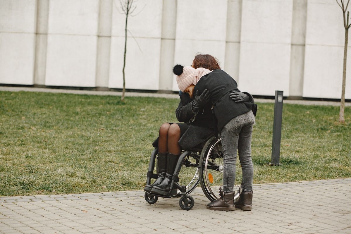 Two people sharing a warm embrace outdoors, one of them seated in a wheelchair