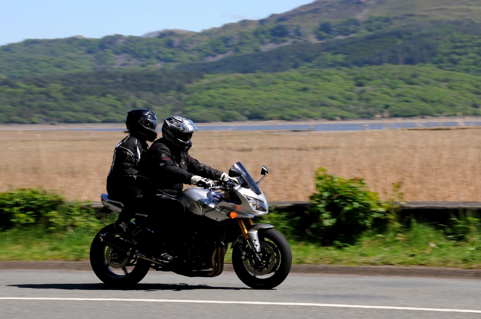 Two riders speeding two-up along a coastal road on a sport motorcycle, passing green hills and a lake