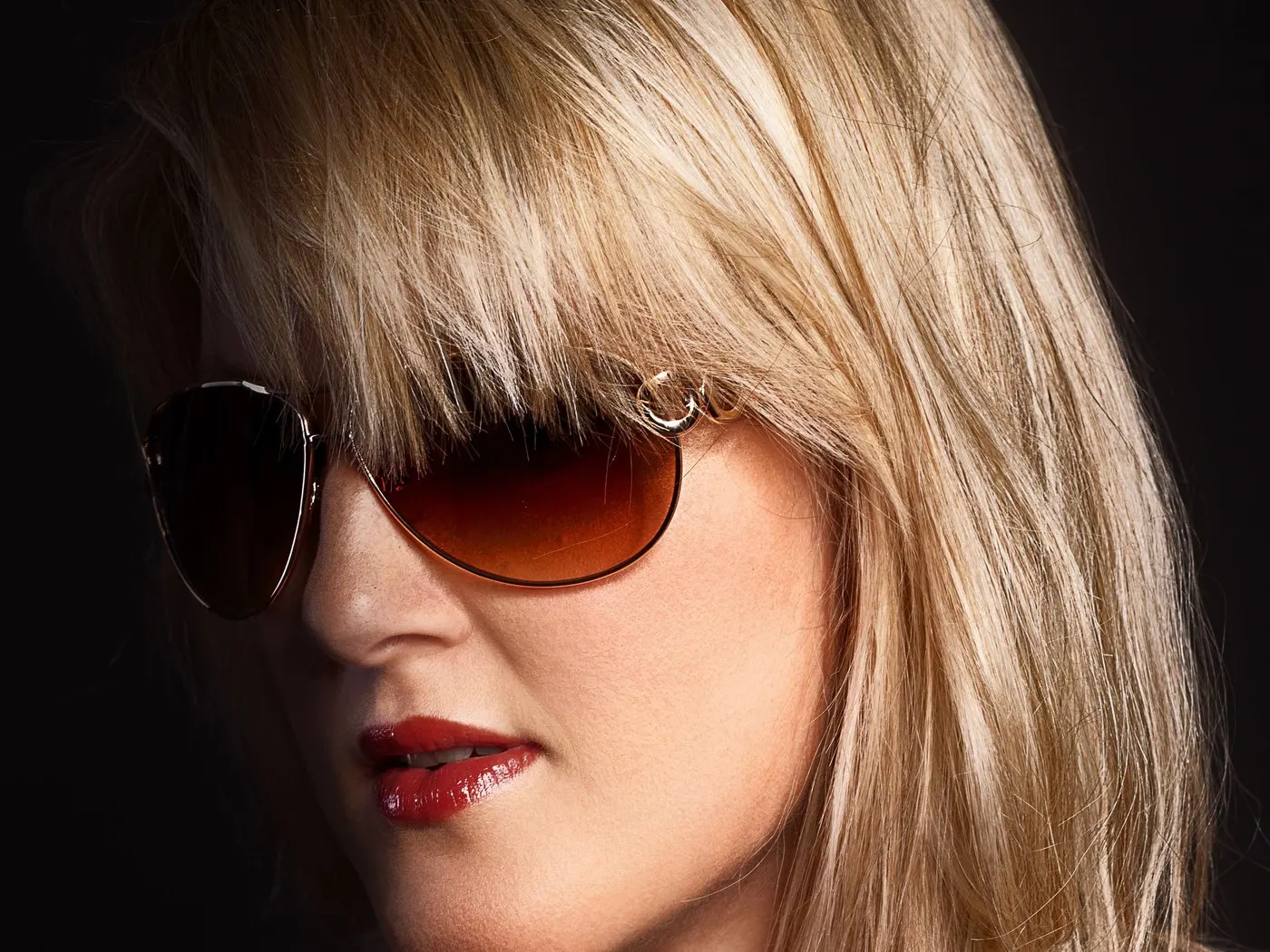 Blonde woman in tinted sunglasses and red lipstick against a dark background