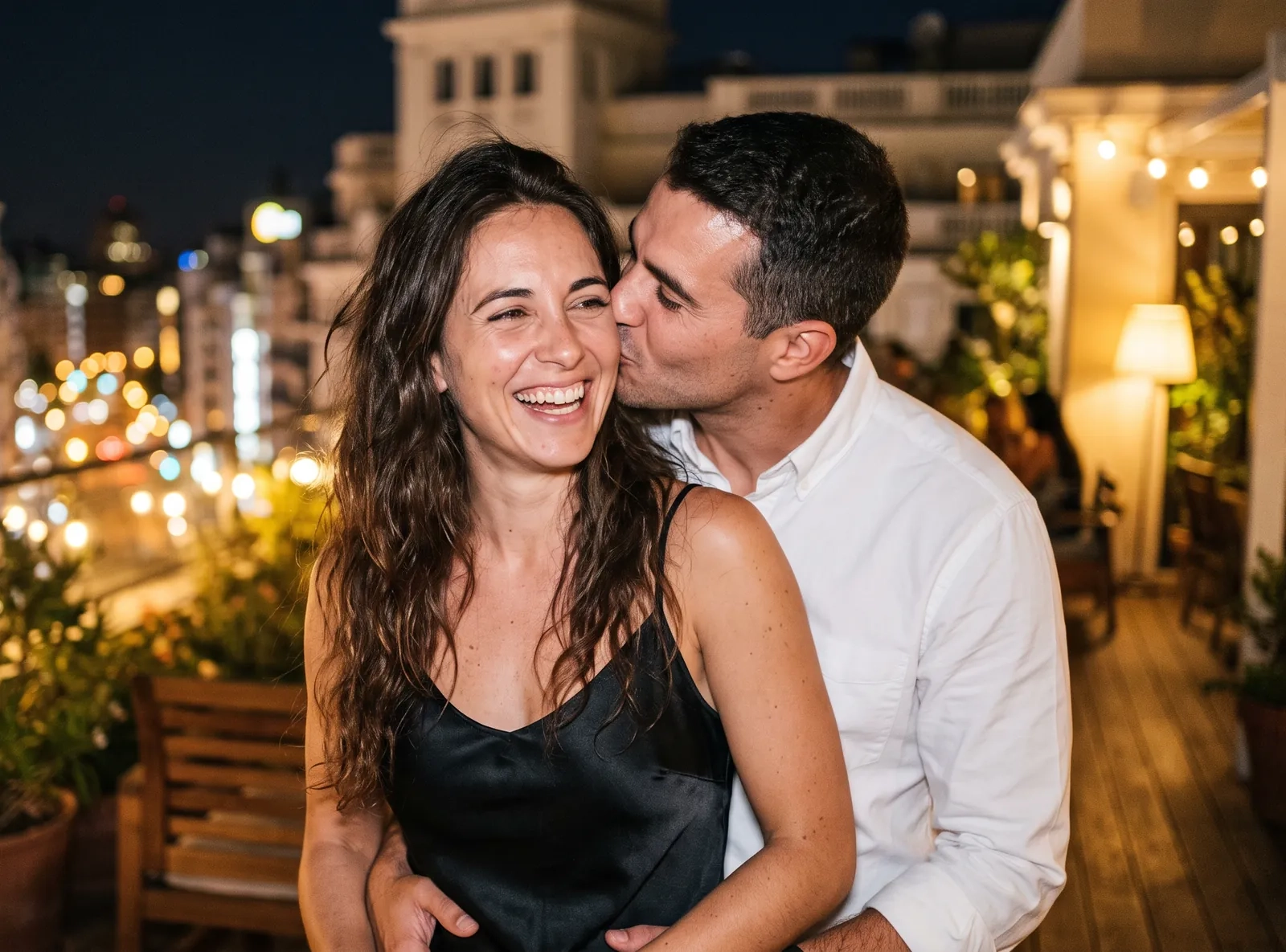 Una mujer con vestido lencero negro se ríe en una azotea con las luces de la ciudad detrás mientras su pareja la abraza por la cintura y le besa la mejilla (imagen generada por IA)