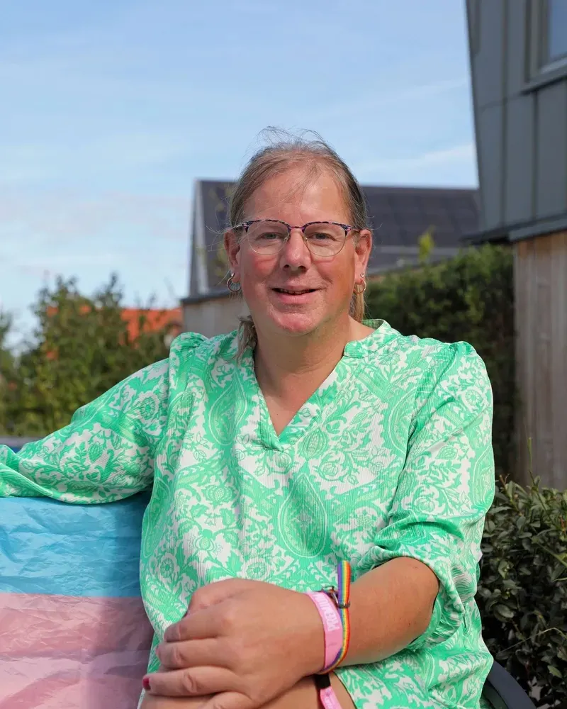 A smiling person in a green patterned top sitting outdoors beside a pink, white and blue flag