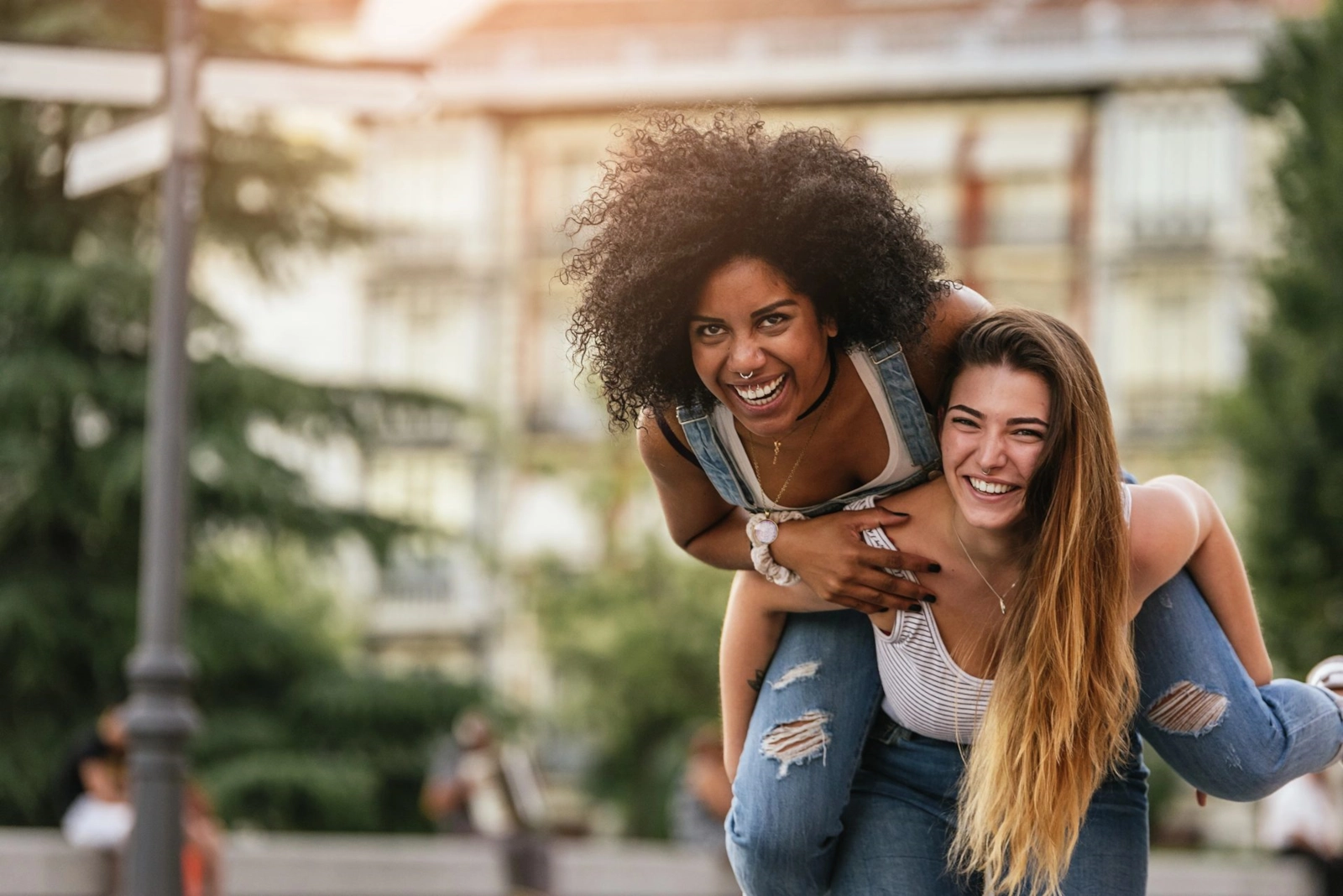 Two women laughing in a city street, one riding piggyback on the other