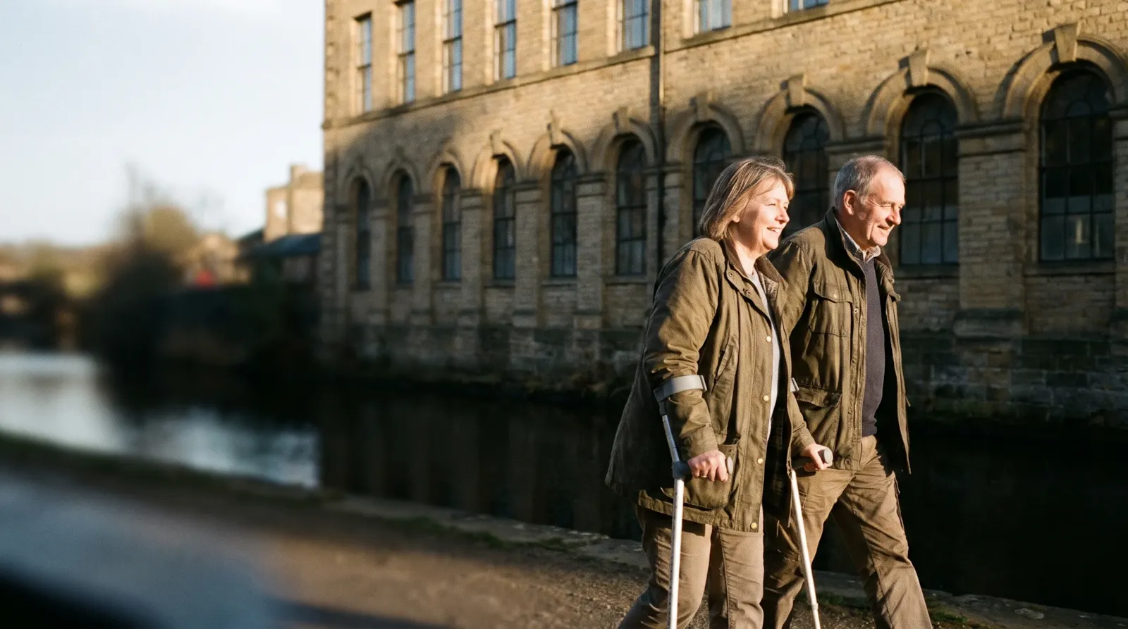 A couple walking beside the canal at Saltaire, the old mill behind them; the woman uses forearm crutches