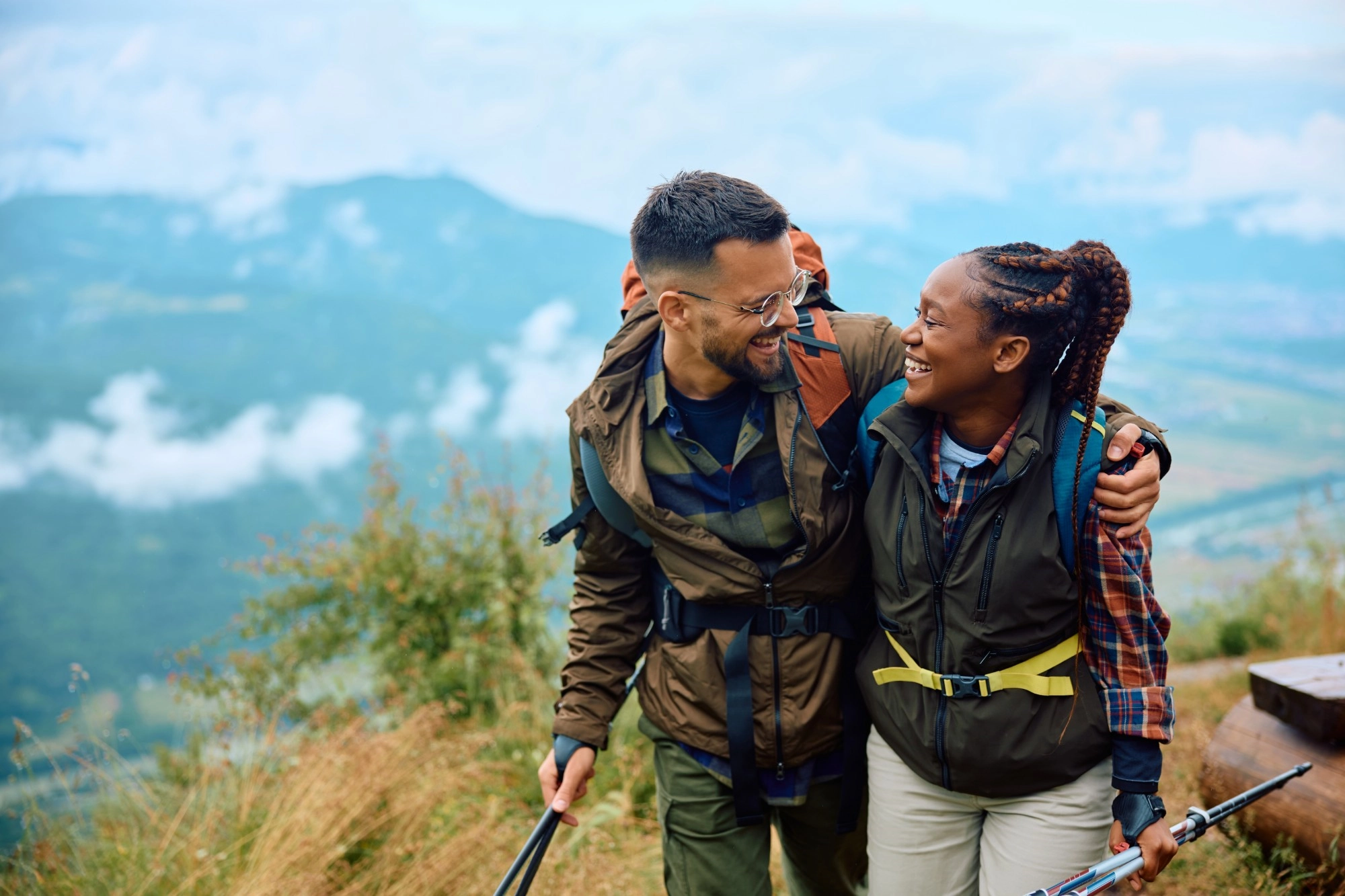 A laughing man and woman with backpacks and walking poles hiking arm in arm above a misty valley