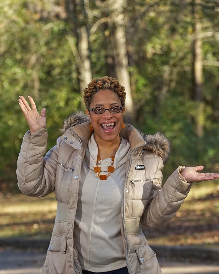 A laughing woman in glasses and a cream puffer jacket with one hand raised, standing in sunny woodland