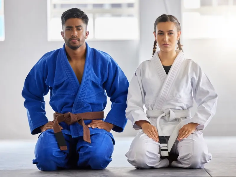 A man in a blue gi and a woman in a white gi kneeling side by side on the mat in a bright dojo