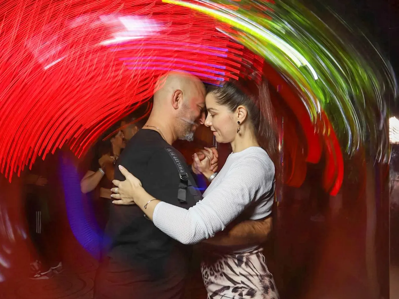 A bald man and a woman in a zebra-print skirt dancing close on a club dance floor, streaks of red and green light around them
