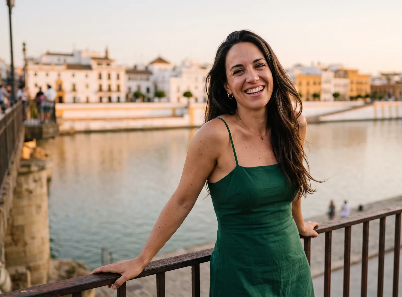 Una mujer morena con vestido verde sonríe apoyada en la barandilla de la calle Betis, en Triana, con el Guadalquivir al atardecer detrás (imagen generada por IA)