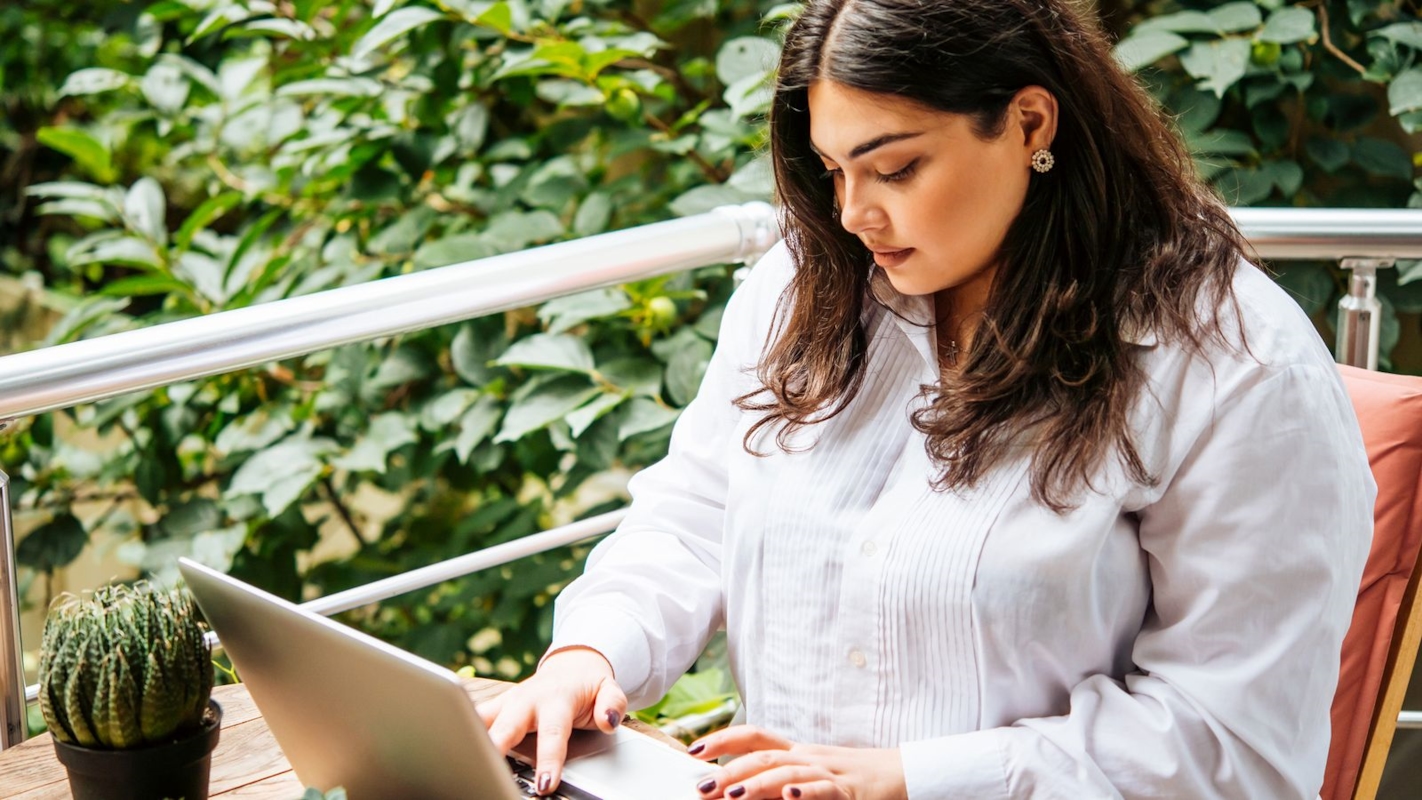 A woman thoughtfully typing on her laptop outdoors on a plant-filled balcony