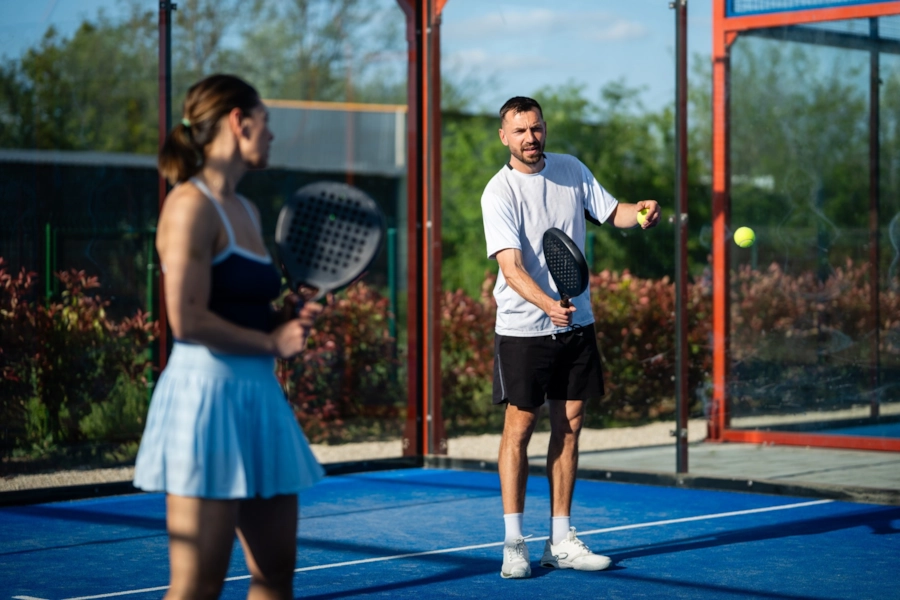 A man and a woman playing mixed doubles on a blue outdoor padel court, the man about to serve