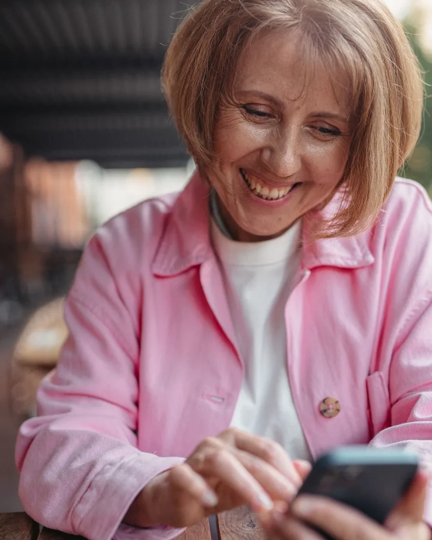 A smiling woman in a pink jacket looking at her phone at an outdoor café table