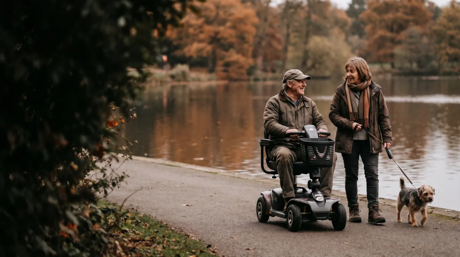 A couple by the lake at Trentham Gardens in autumn; the man rides a mobility scooter and the woman walks their dog