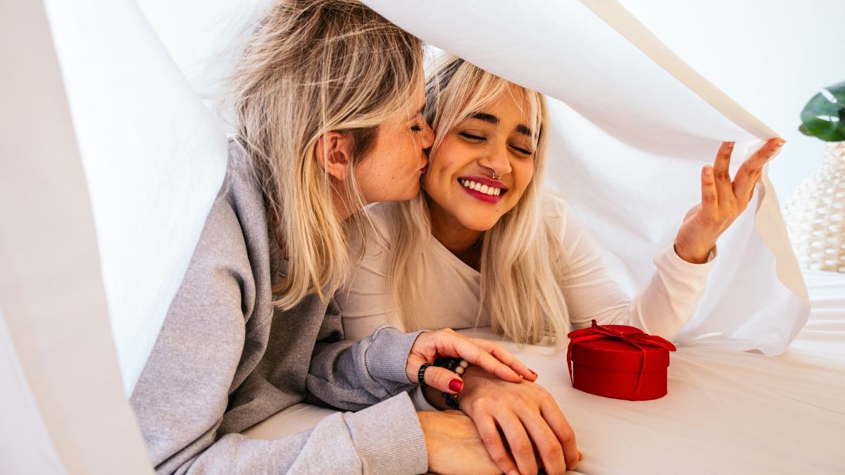 Two women laughing together at home under a sheet, one kissing the other on the cheek