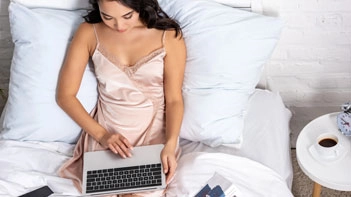Woman in a champagne satin camisole sitting on a bed with a laptop and a cup of coffee