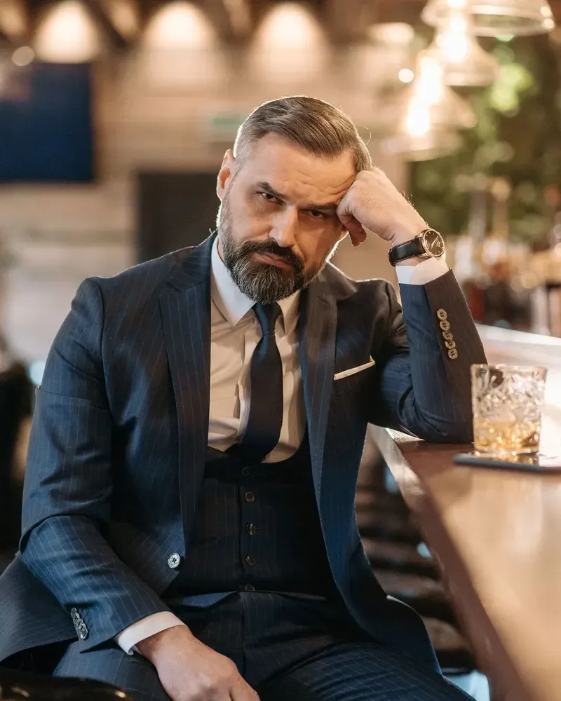 A man with a greying beard in a pinstripe three-piece suit sitting at a bar with a whisky, giving the camera a direct look