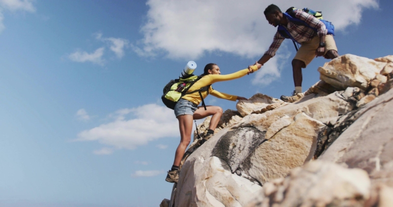 A man reaching down to help a woman up a rocky outcrop under a blue sky