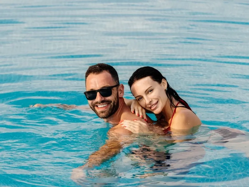 A smiling couple in sunglasses with their arms around each other in a sparkling blue pool