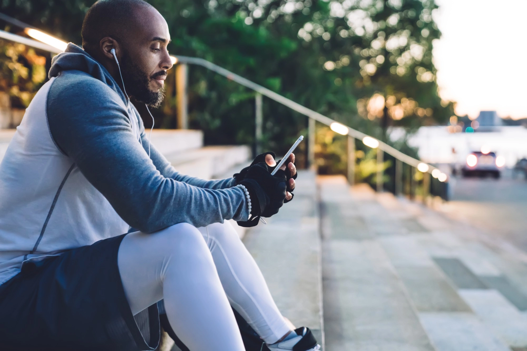 A man in workout clothes and earphones sitting on outdoor steps looking at his phone