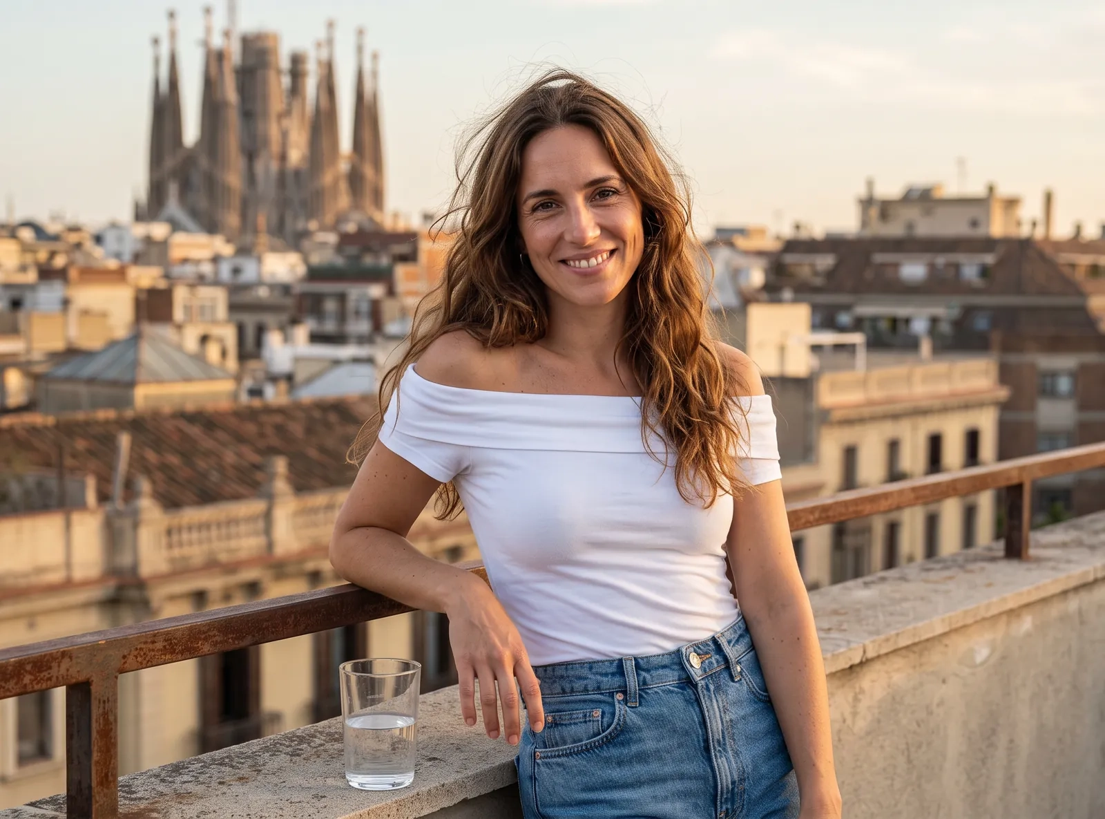 Una mujer sonriente de pelo castaño ondulado y top blanco con los hombros al aire se apoya en la barandilla de una azotea de Barcelona al atardecer, con la Sagrada Familia al fondo (imagen generada por IA)