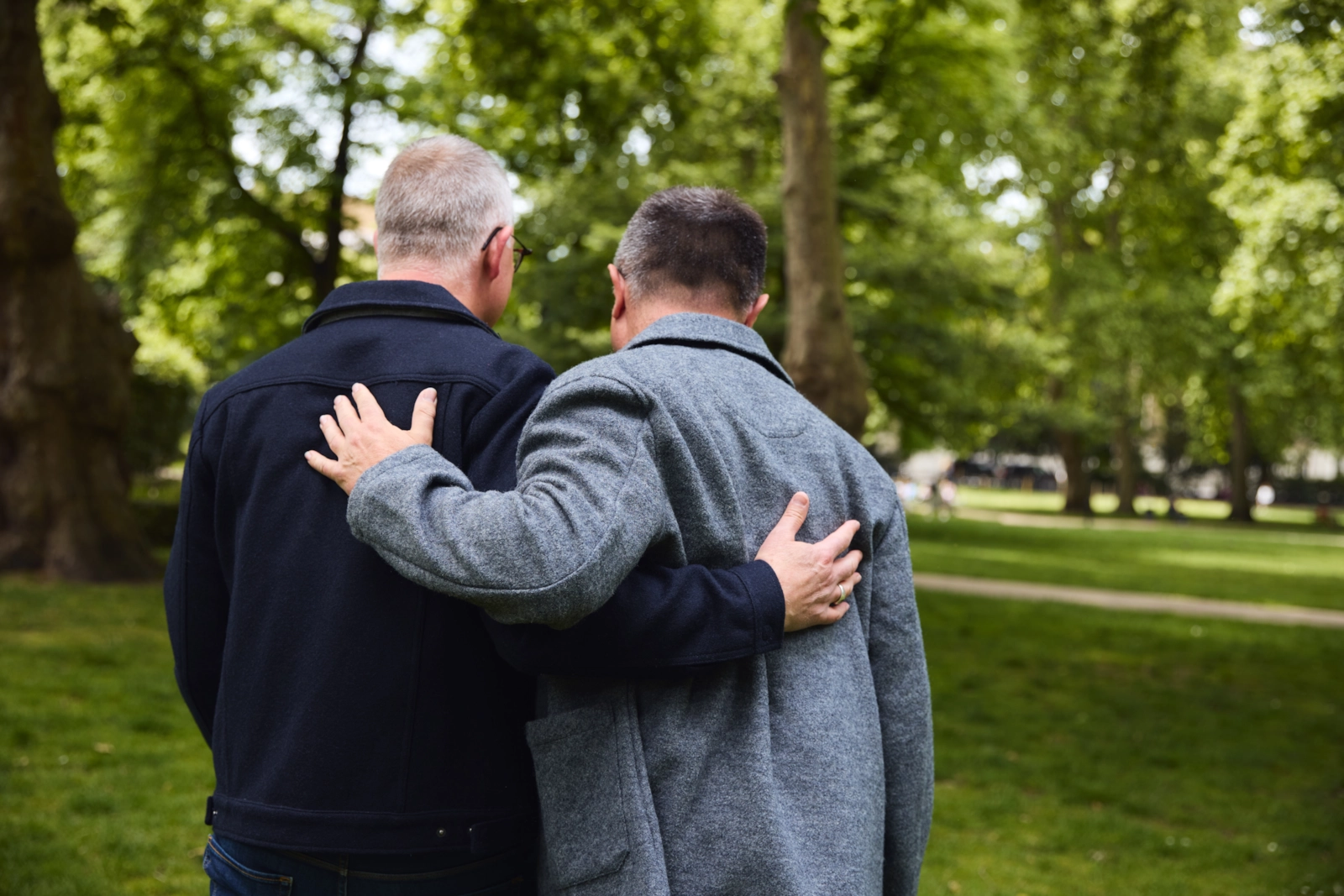 Dos hombres mayores paseando por un parque, uno con el brazo sobre el hombro del otro.
