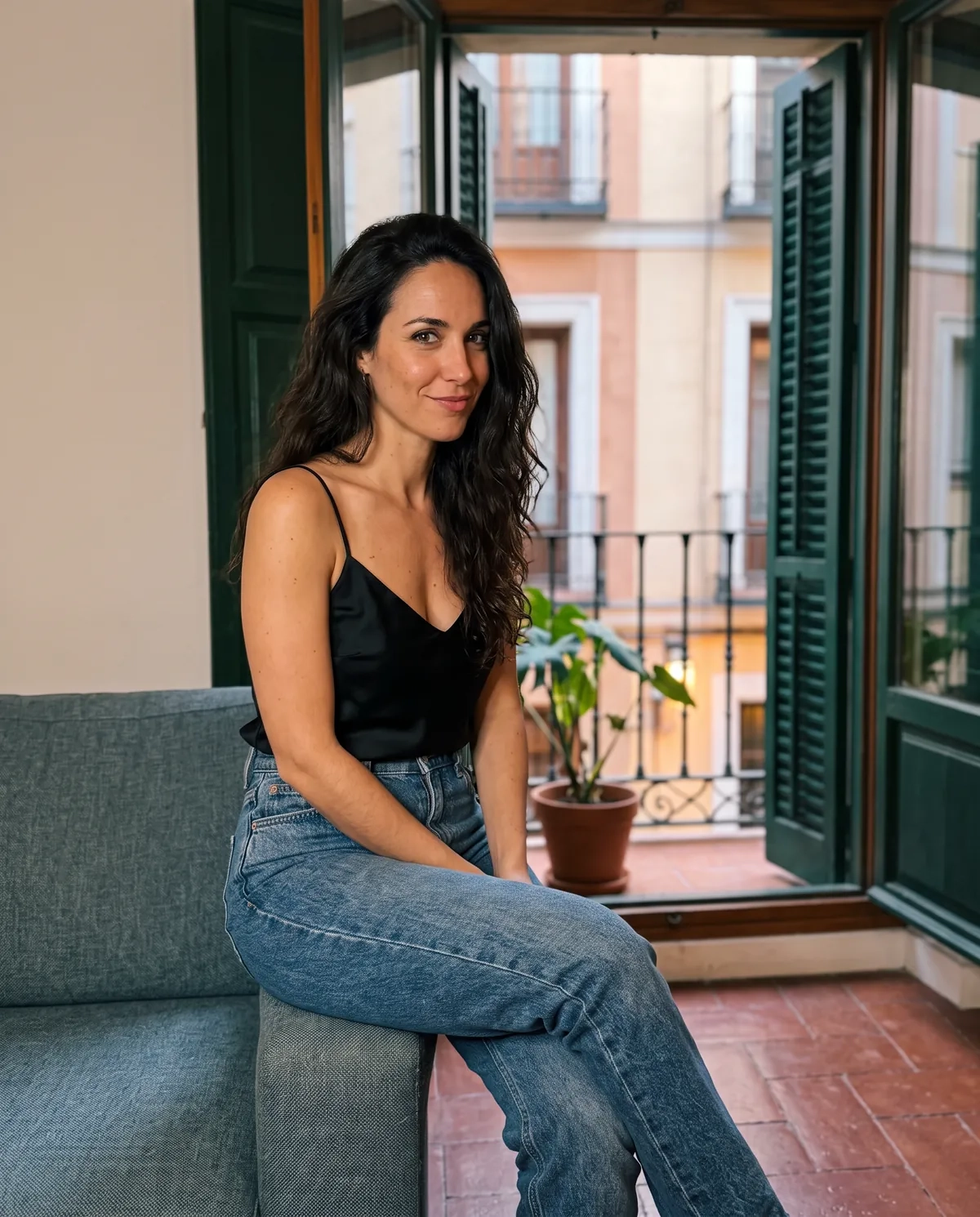 Una mujer de pelo largo, oscuro y ondulado, con un top negro de tirantes y vaqueros, sentada en el brazo de un sofá junto a un balcón abierto de un piso de Madrid, sonríe a la cámara (imagen generada por IA)