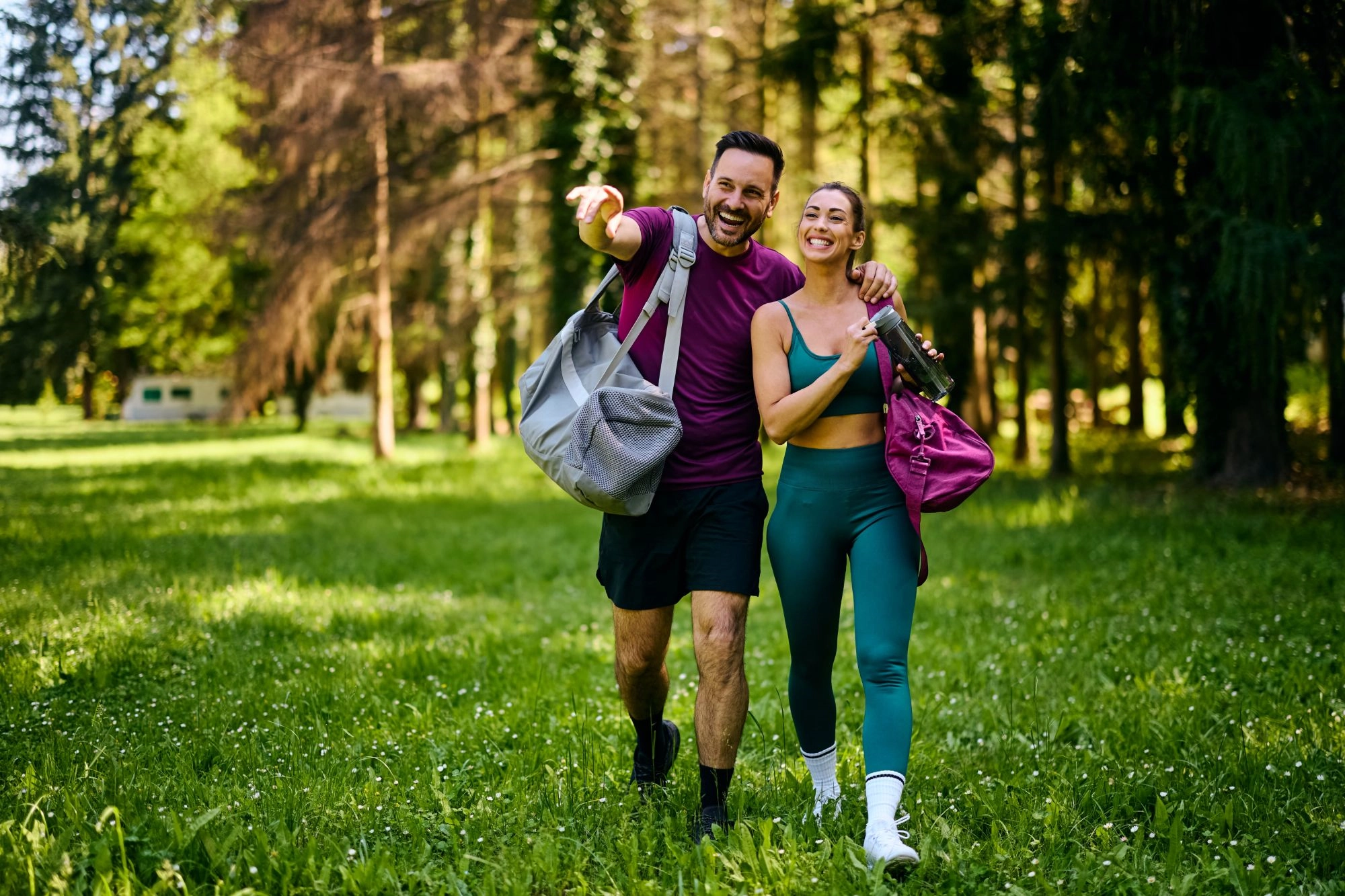 A laughing man and woman with gym bags walking arm in arm across a sunny park meadow