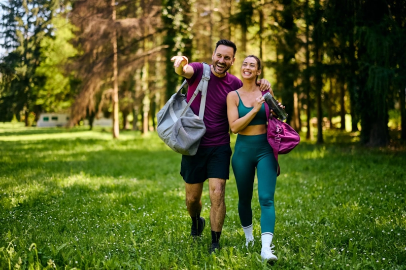 A laughing man and woman with gym bags walking arm in arm across a sunny park meadow