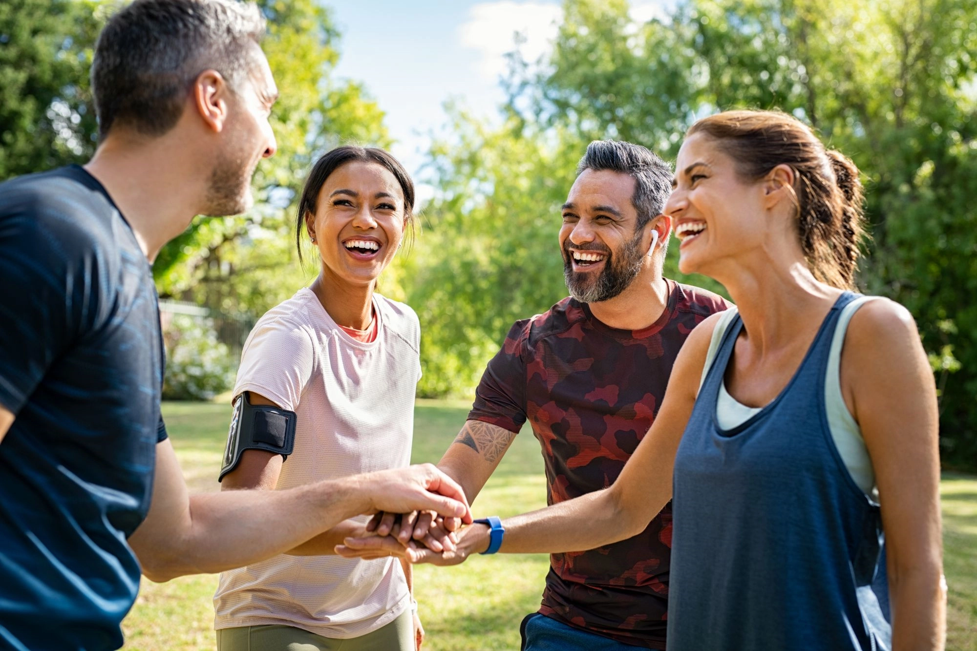 Four laughing friends in workout clothes stacking their hands together in a sunny park