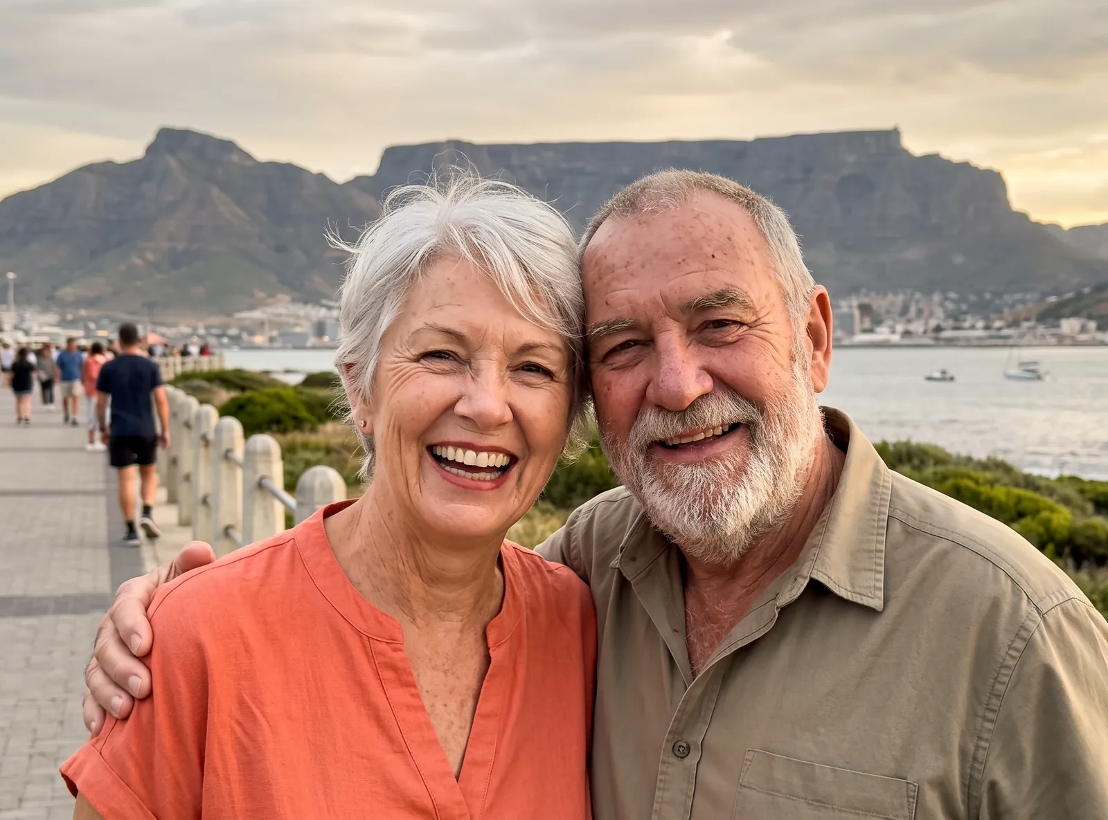 A couple in their sixties laugh together on a seaside promenade with a flat-topped mountain behind them, she in a coral blouse (AI-generated image)