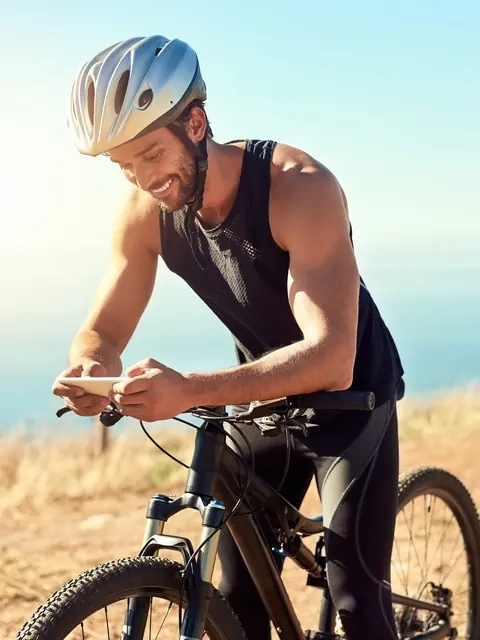 A smiling cyclist in a helmet checking his phone on a coastal trail