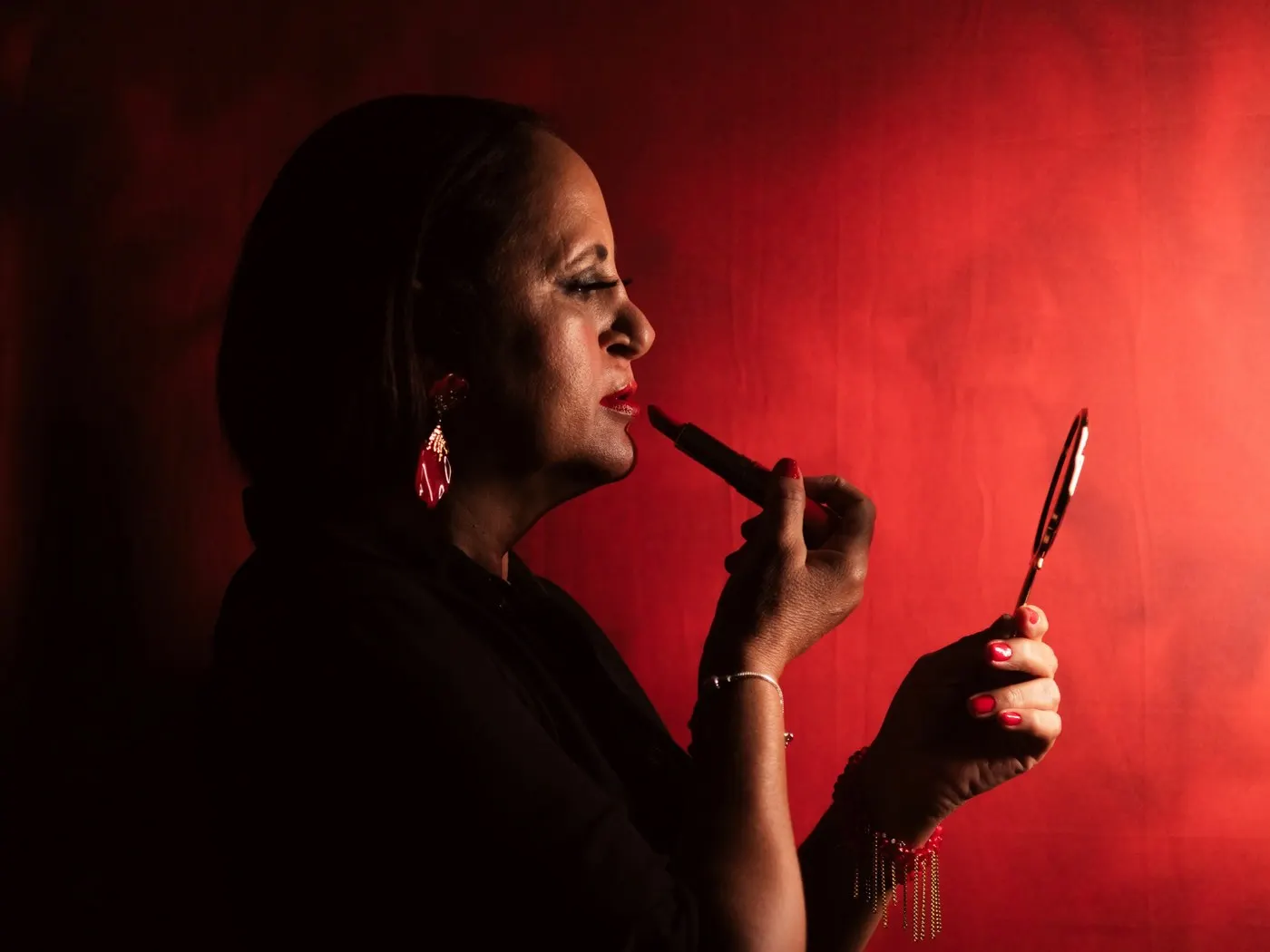 A woman in black, side-on against a red backdrop, putting on red lipstick in a small hand mirror