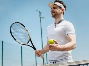 A smiling tennis player in a white polo holding his racket and a ball