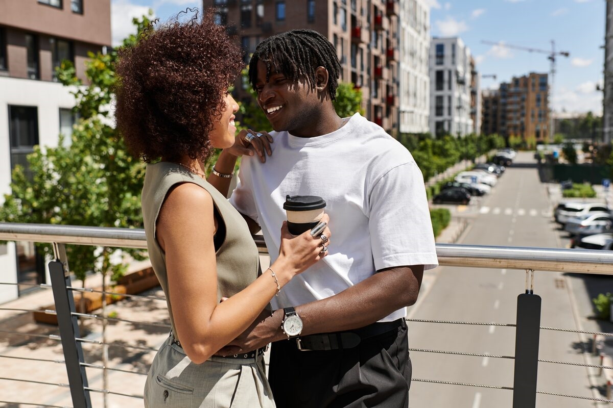 A couple sharing coffee together at an outdoor spot in the city, smiling and clearly comfortable in each other's company