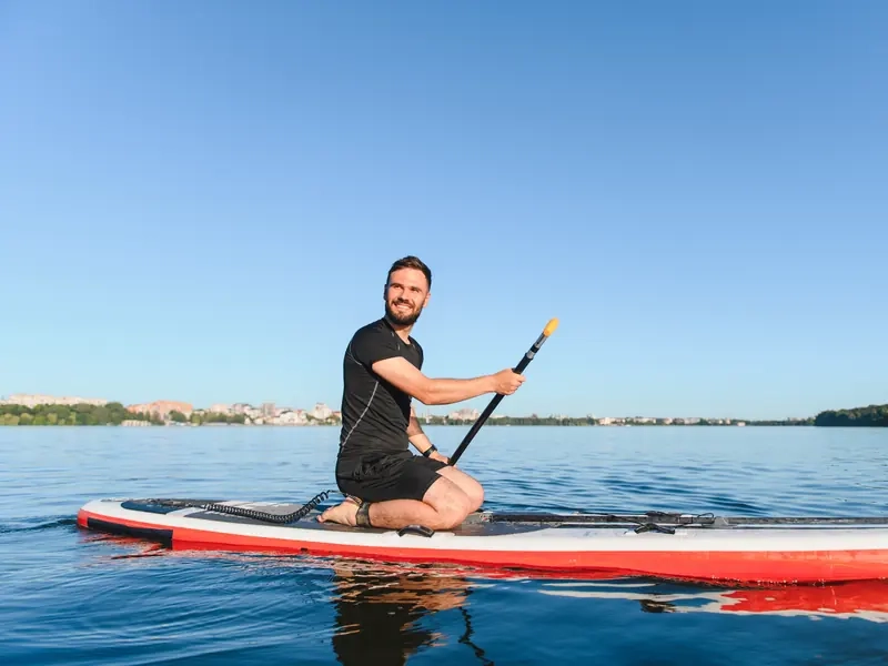 A smiling man kneeling on a red paddleboard on a calm blue lake