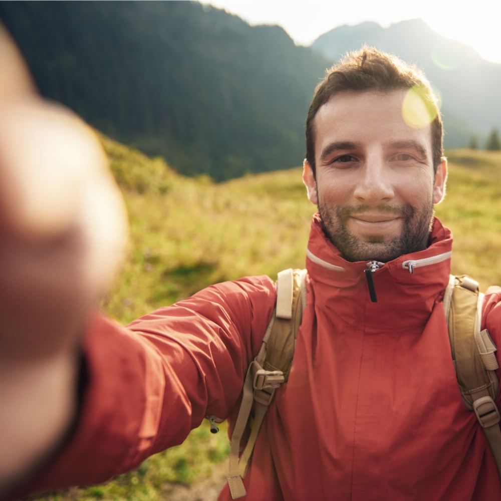 Un hombre con chaqueta roja y mochila sonríe mientras se toma una selfie en la montaña