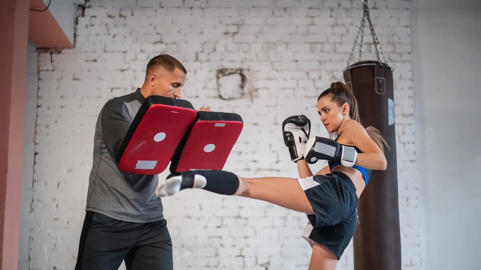 A woman in boxing gloves landing a high kick on pads held by her training partner in a brick-walled gym