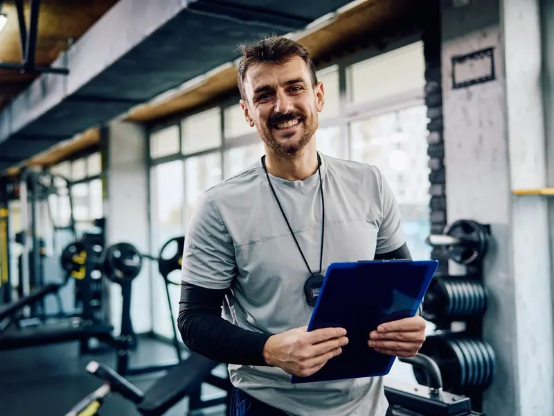 A smiling male personal trainer holding a clipboard in a gym