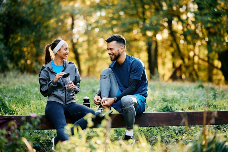 A laughing woman and man taking a break on a wooden rail in a sunny park, one tying his running shoe