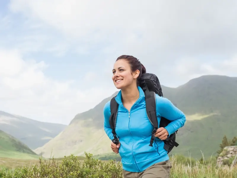 A smiling woman with a backpack walking a mountain trail