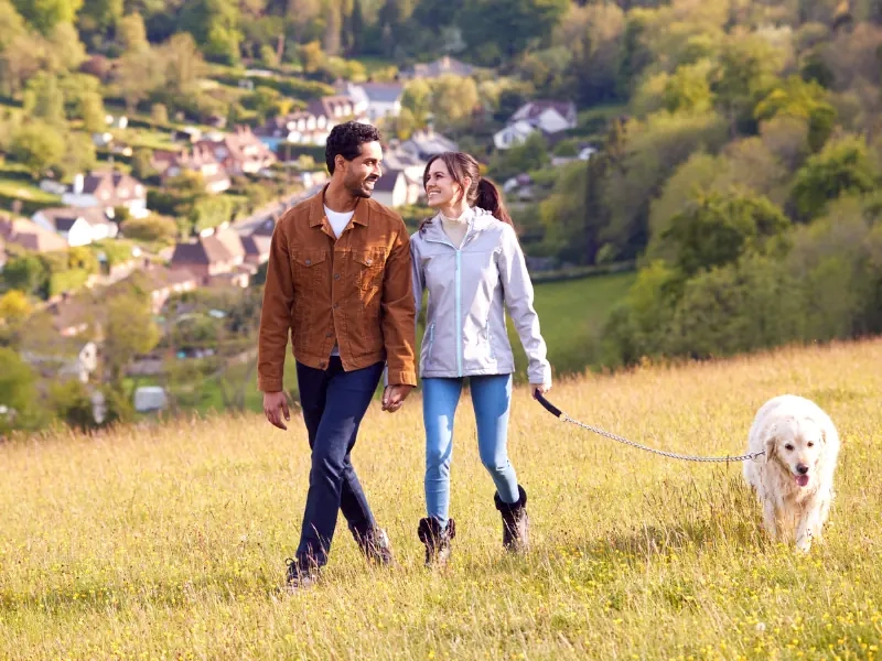 A couple walking a white dog across a hillside above a village in the valley
