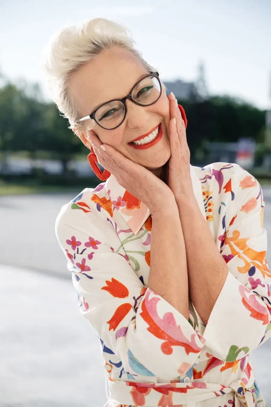 A laughing woman with short grey hair and glasses, hands on her cheeks, in a bright floral shirt