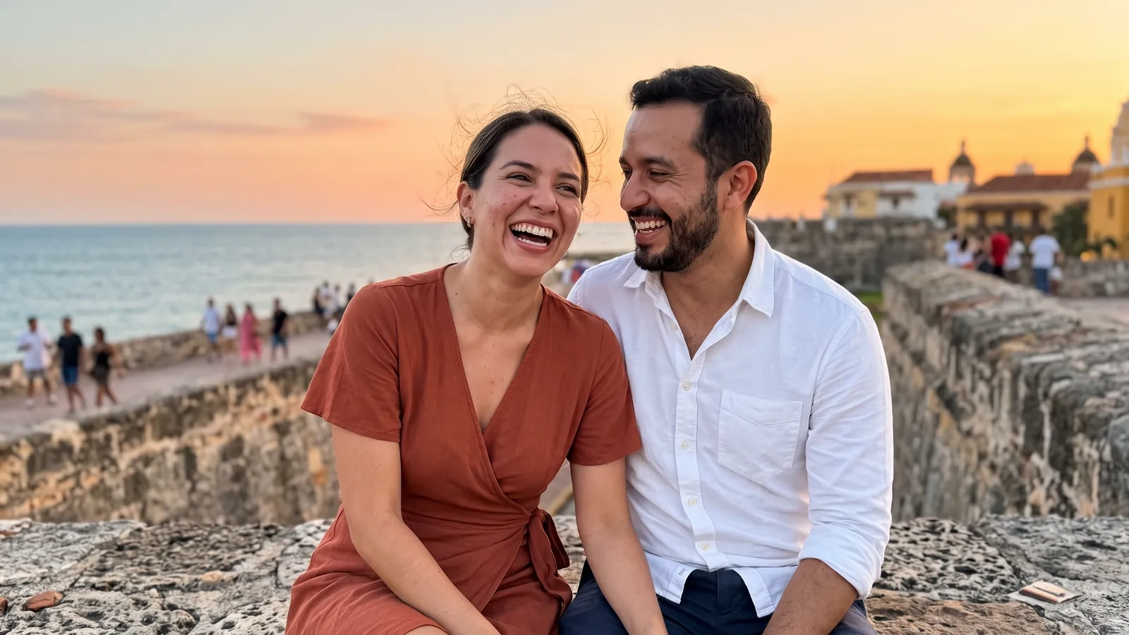 Una pareja se ríe sentada muy cerca sobre la muralla de Cartagena al atardecer, con el mar Caribe y un cielo naranja detrás (imagen generada por IA)