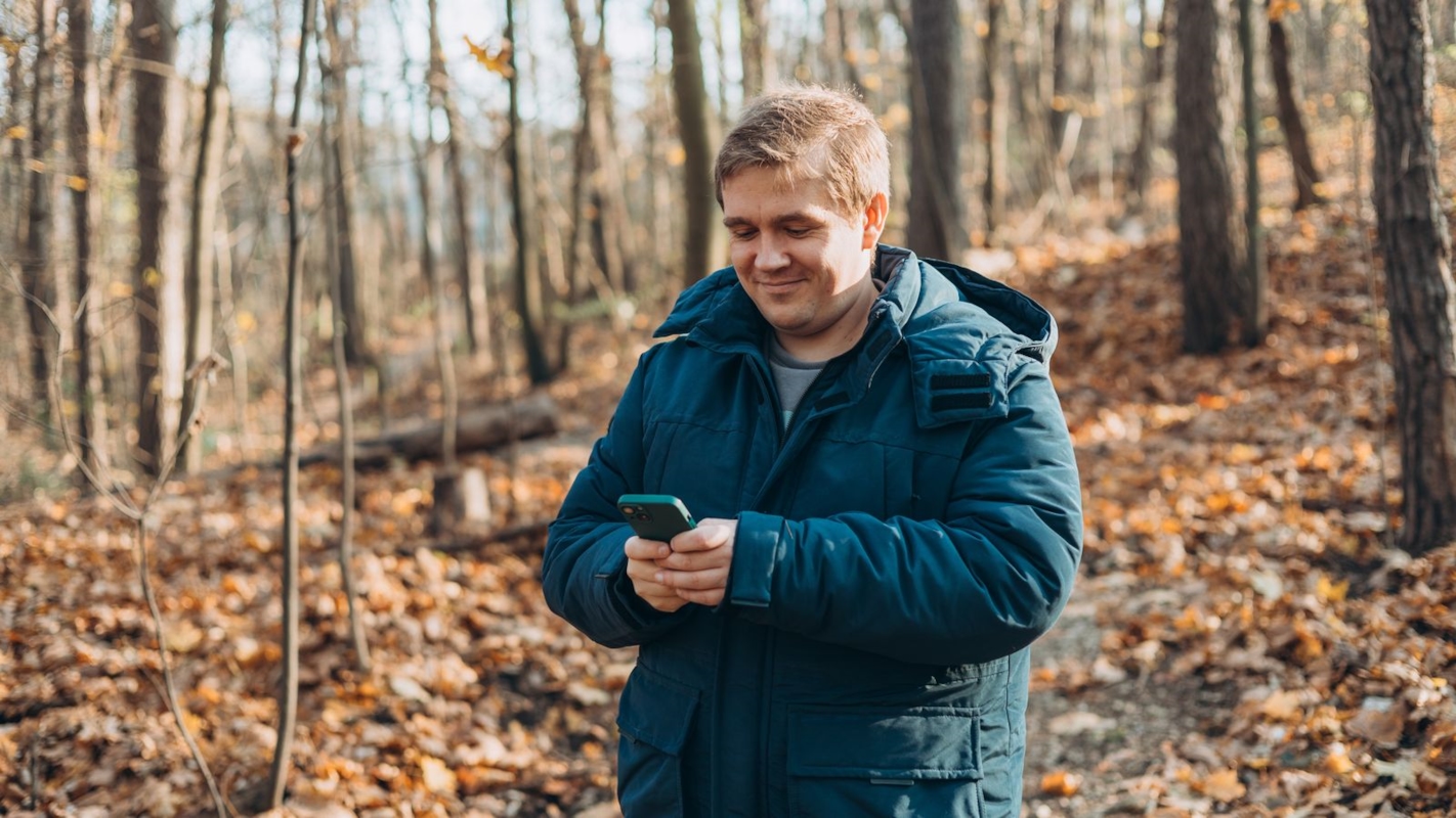 A man smiling at his phone while walking through an autumn forest